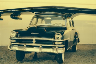A vintage, shiny black car is displayed under an aircraft wing, giving it an unusual hybrid appearance. The car is well-preserved with a classic design featuring chrome accents and prominent headlights.