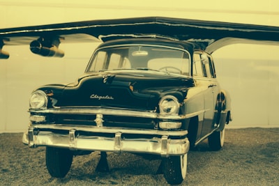 A vintage, shiny black car is displayed under an aircraft wing, giving it an unusual hybrid appearance. The car is well-preserved with a classic design featuring chrome accents and prominent headlights.