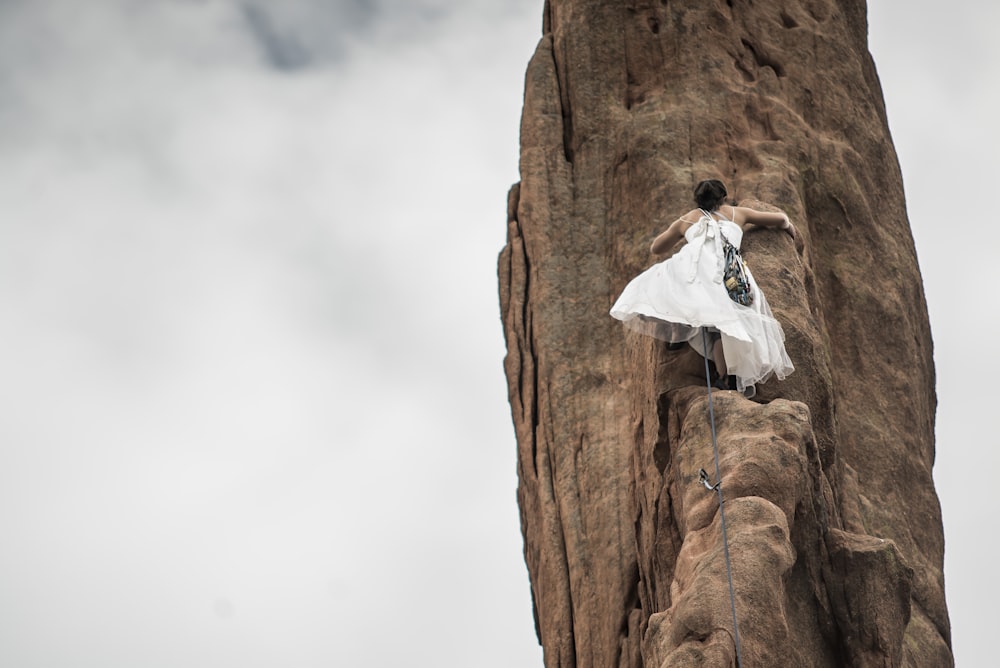 白いウェディングドレスを着た女性が白い空の下で茶色の岩に登る