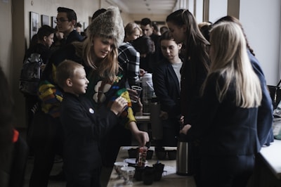 A group of people, including children and adults, are gathered indoors around tables with various items and beverages. The atmosphere seems interactive as a woman wearing a colorful sweater and fur hat engages with a young boy, possibly teaching or sharing something with him. Other individuals in the background are involved in their own activities or conversations, creating a lively environment.