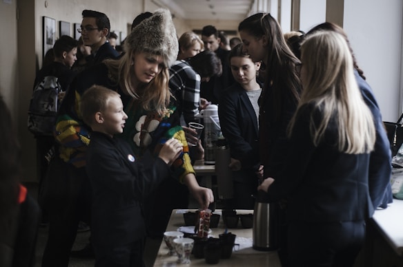 A group of people, including children and adults, are gathered indoors around tables with various items and beverages. The atmosphere seems interactive as a woman wearing a colorful sweater and fur hat engages with a young boy, possibly teaching or sharing something with him. Other individuals in the background are involved in their own activities or conversations, creating a lively environment.