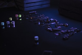 A dimly lit scene features a collection of miscellaneous metal objects, possibly keys and small tools, spread out on a dark surface. Neon-colored containers, including pink, yellow, and blue, are placed to the side. A wooden crate rests in the background, adding to the shadowy ambiance.