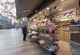 A vibrant market stall filled with an array of colorful sweets, chocolates, and souvenirs. Shelves are stocked with packaged candies, lollipops, and beautifully wrapped confections. An elderly person stands in front, examining the neatly arranged items. The background reveals a bustling pedestrian street with people walking along, and trees lining the path.