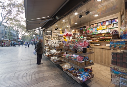A vibrant market stall filled with an array of colorful sweets, chocolates, and souvenirs. Shelves are stocked with packaged candies, lollipops, and beautifully wrapped confections. An elderly person stands in front, examining the neatly arranged items. The background reveals a bustling pedestrian street with people walking along, and trees lining the path.