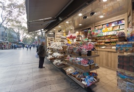 A vibrant market stall filled with an array of colorful sweets, chocolates, and souvenirs. Shelves are stocked with packaged candies, lollipops, and beautifully wrapped confections. An elderly person stands in front, examining the neatly arranged items. The background reveals a bustling pedestrian street with people walking along, and trees lining the path.