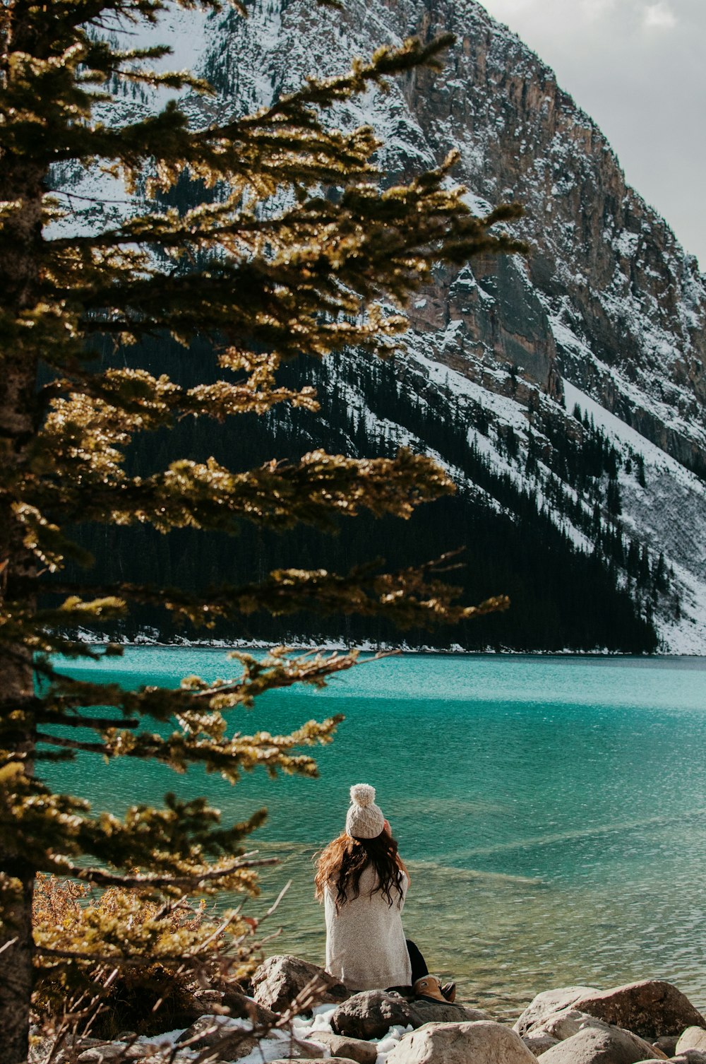 woman sitting on stone facing body of water and glacier mountain during day