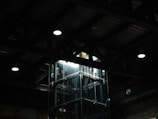 A dimly lit industrial interior with a glass elevator shaft. Overhead lighting casts minimal illumination, highlighting the metal beams and ceiling structure.