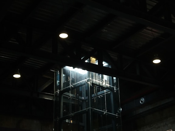 A dimly lit industrial interior with a glass elevator shaft. Overhead lighting casts minimal illumination, highlighting the metal beams and ceiling structure.
