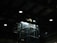 A dimly lit industrial interior with a glass elevator shaft. Overhead lighting casts minimal illumination, highlighting the metal beams and ceiling structure.