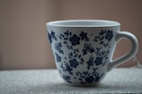 A white ceramic mug with a blue floral pattern sits on a textured surface. The design includes various flowers and leaves in a symmetrical arrangement around the cup.
