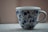 A white ceramic mug with a blue floral pattern sits on a textured surface. The design includes various flowers and leaves in a symmetrical arrangement around the cup.