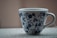 A white ceramic mug with a blue floral pattern sits on a textured surface. The design includes various flowers and leaves in a symmetrical arrangement around the cup.