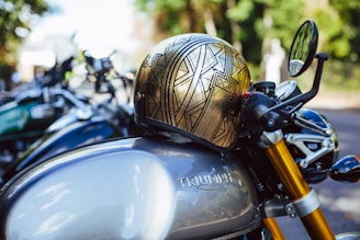A detailed view of a Triumph motorcycle parked outdoors with a metallic helmet placed on the tank. The helmet features an intricate geometric design with the letters 'DGR'. The motorcycle's sleek design includes golden front forks and a side mirror that is slightly blurred in the background. Other motorcycles can be seen out of focus behind it, suggesting a gathering or event.