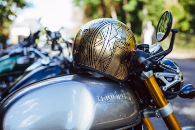 A detailed view of a Triumph motorcycle parked outdoors with a metallic helmet placed on the tank. The helmet features an intricate geometric design with the letters 'DGR'. The motorcycle's sleek design includes golden front forks and a side mirror that is slightly blurred in the background. Other motorcycles can be seen out of focus behind it, suggesting a gathering or event.