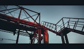 A steel framework of industrial piping and walkways, silhouetted against a pale sky. The structure features latticework and railings, highlighted by a warm, reddish glow from the setting sun.