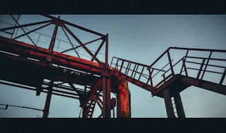 A steel framework of industrial piping and walkways, silhouetted against a pale sky. The structure features latticework and railings, highlighted by a warm, reddish glow from the setting sun.