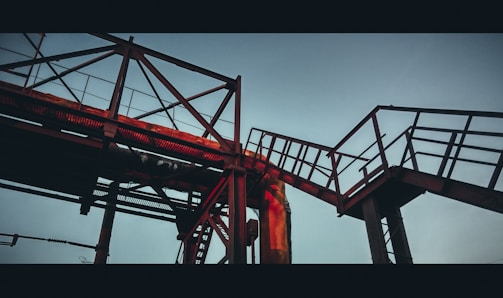 A steel framework of industrial piping and walkways, silhouetted against a pale sky. The structure features latticework and railings, highlighted by a warm, reddish glow from the setting sun.
