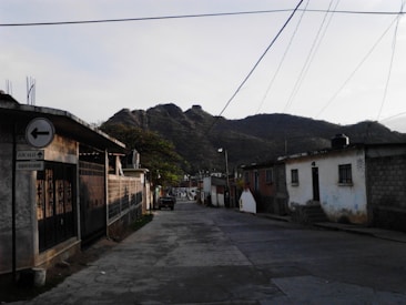 A quiet street scene with modest houses lining the road and a mountainous backdrop. Power lines stretch across the sky and a sign points towards a direction labeled 'Zocalo'. The lighting suggests early morning or late afternoon, casting soft shadows.