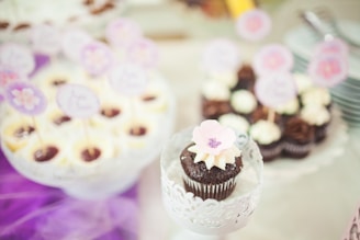 A beautifully arranged display of cupcakes, with a chocolate cupcake in the foreground adorned with a pale pink flower decoration. In the background, there are more cupcakes topped with cream and small decorative toppers. The setting has a soft pastel ambiance with purple accents.