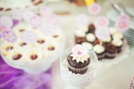 A beautifully arranged display of cupcakes, with a chocolate cupcake in the foreground adorned with a pale pink flower decoration. In the background, there are more cupcakes topped with cream and small decorative toppers. The setting has a soft pastel ambiance with purple accents.