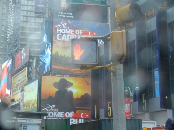 A busy urban street scene with multiple large billboards and advertisements, including a prominent ad for Turkey featuring Capadoccia and Istanbul. There is a pedestrian crossing signal showing a red hand, indicating not to walk. The scene is slightly blurred, possibly from rain or fog.