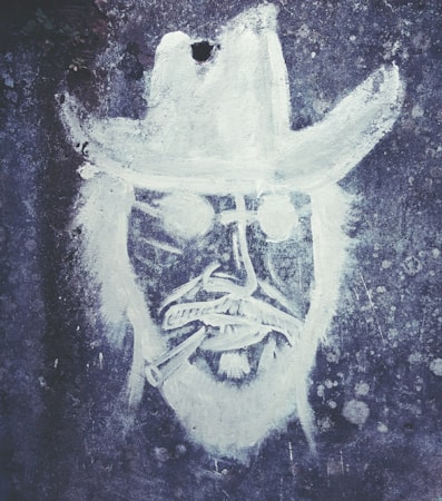 A monochromatic graffiti drawing on a textured wall depicting a person's face wearing a wide-brimmed hat and sunglasses, with a cigarette in their mouth. The art style is rough and sketch-like, focusing on bold outlines and minimal detail.