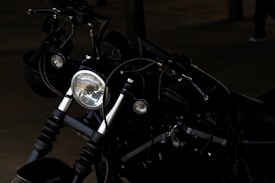 A close-up of a motorcycle's front, highlighting the headlight and handlebars. The motorcycle appears to be parked in a dimly lit area, and its dark color is prominent. There are visible wires and mechanical parts around the headlight.
