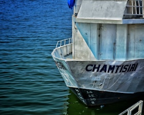 The image features the rear view of a large, white boat named 'Chamtsiri' docked on a calm body of water. The metal surfaces of the boat reflect sunlight, showing some signs of rust and aging. The water appears deep blue and gently ripples around the vessel.