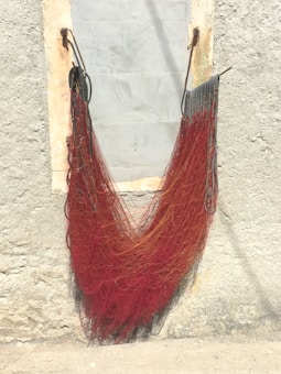 A bundle of red fishing nets is hanging on a weathered, textured wall with hooks on both sides. The wall is light gray with patches of peeling paint, creating a rustic atmosphere.