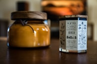 Two candles placed on a wooden surface, one in an amber glass jar with a wooden lid and the other in a clear glass container with a vintage-style label featuring ornate typography and illustrations.