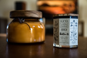 Two candles placed on a wooden surface, one in an amber glass jar with a wooden lid and the other in a clear glass container with a vintage-style label featuring ornate typography and illustrations.