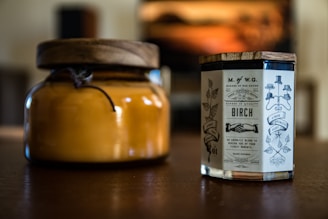 Two candles placed on a wooden surface, one in an amber glass jar with a wooden lid and the other in a clear glass container with a vintage-style label featuring ornate typography and illustrations.