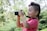 A young child with a mohawk hairstyle holds a camera, enthusiastically taking a photograph. The child is wearing a pink shirt and is surrounded by lush greenery, suggesting a natural outdoor setting.