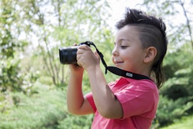 A young child with a mohawk hairstyle holds a camera, enthusiastically taking a photograph. The child is wearing a pink shirt and is surrounded by lush greenery, suggesting a natural outdoor setting.