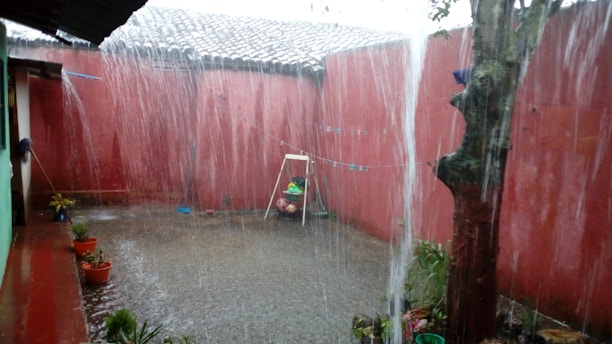 The image captures a heavily raining scene in an enclosed courtyard. Rainwater is pouring heavily from the roof, creating a thick curtain of water. The ground is wet and filled with puddles. There is a simple swing set with a child-sized seat in the background. Potted plants are lined up next to the walls, which are painted red. A large tree is visible on the right side.