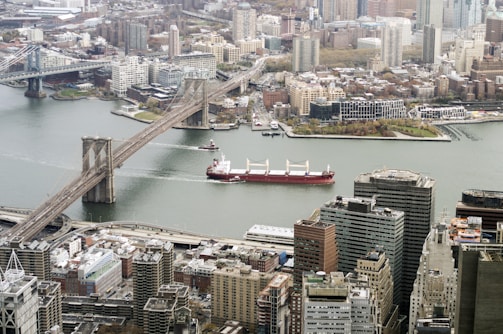 An aerial view of a cityscape featuring a prominent bridge spanning a river. A large cargo ship is sailing beneath the bridge. The surrounding city is filled with high-rise buildings and a mix of modern and older architecture. The riverbanks are lined with greenery and urban developments.
