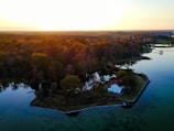 An aerial view of a small island with a white house featuring a red roof near the water's edge. The island is surrounded by a calm body of water reflecting the sky, with dense trees that display autumn colors. In the background, the sun is setting, casting a golden hue over the landscape.