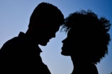Two people facing each other in silhouette, with a clear blue sky in the background. The figures are positioned closely, suggesting an intimate or affectionate relationship.