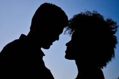 Two people facing each other in silhouette, with a clear blue sky in the background. The figures are positioned closely, suggesting an intimate or affectionate relationship.