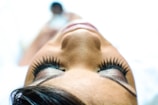 A close-up view of a person lying down with focus on the eyes, which are adorned with long, elegant lashes. The image captures the serene and calm expression on the person's face, enhanced by a soft focus and bright lighting.