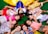 A person dressed in a Snow White costume is lying on the grass surrounded by a group of children, all lying in a circle with their heads together. The children are wearing bright and colorful clothes, some with floral and striped patterns, and they all appear to be smiling and having fun.