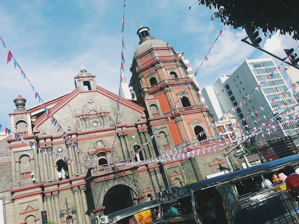 A historic church with ornate architecture stands prominently, featuring a facade with bell towers. Multicolored triangular flags are strung across the scene, adding a festive ambiance. A jeepney is visible in the foreground with people inside. Modern buildings in the background enhance the contrast between tradition and modernity.