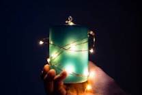 A hand gently holds a large teal candle wrapped with delicate, glowing string lights. The background is dark, creating a contrast that emphasizes the soft illumination of the lights and the tranquil hue of the candle.