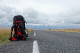 A large hiking backpack rests on the side of a long, empty road that stretches into the distance. The sky is overcast with thick, gray clouds, and golden fields of grass line the road on either side. The setting conveys a sense of solitude and adventure.
