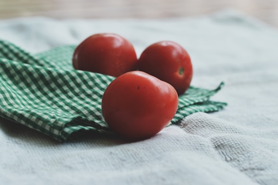 Three ripe, red tomatoes are placed on a green and white checkered cloth, which lies on a textured, light-colored surface. The image is softly focused, creating a rustic and homely feel.