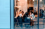 A bustling cafe scene with people sitting at tables, working on laptops and engaging in conversation. The interior features warm lighting and a visible textured wall, creating a cozy atmosphere. Through the large glass windows, a few individuals can be seen standing and talking outside.
