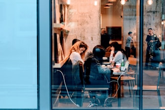 A bustling cafe scene with people sitting at tables, working on laptops and engaging in conversation. The interior features warm lighting and a visible textured wall, creating a cozy atmosphere. Through the large glass windows, a few individuals can be seen standing and talking outside.