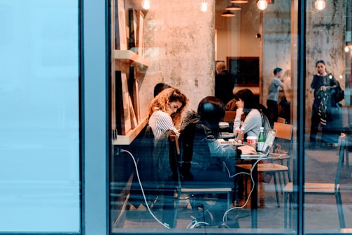 A bustling cafe scene with people sitting at tables, working on laptops and engaging in conversation. The interior features warm lighting and a visible textured wall, creating a cozy atmosphere. Through the large glass windows, a few individuals can be seen standing and talking outside.