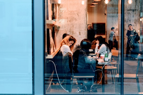 A bustling cafe scene with people sitting at tables, working on laptops and engaging in conversation. The interior features warm lighting and a visible textured wall, creating a cozy atmosphere. Through the large glass windows, a few individuals can be seen standing and talking outside.