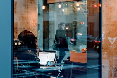 A person is seated at a table inside a cafe, working on a laptop with documents open on the screen. Nearby, a smartphone is propped up, perhaps for video calls. The window reflects a person walking outside and the urban street scene with parked cars.